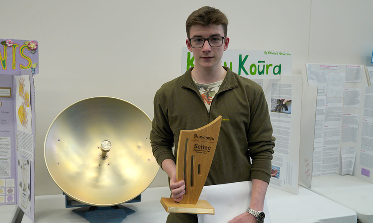 Teenage boy holding a trophy in front of science boards and a science instrument.