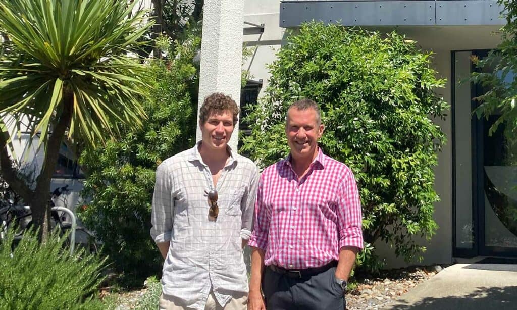 Two men standing in front of trees and a building.