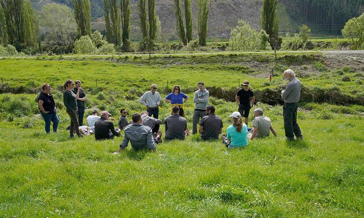 Man speaks to group of people on grassy river bank