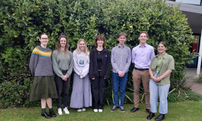 seven students standing in a line in front of trees